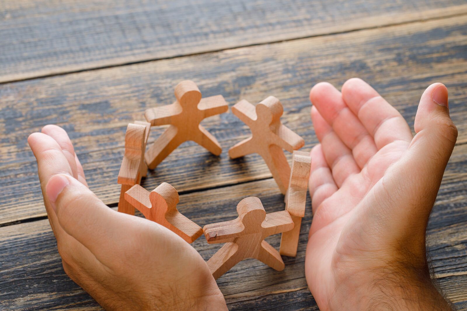 Two cupped hands enclosing six wooden figures holding hands in a circle on a wooden table