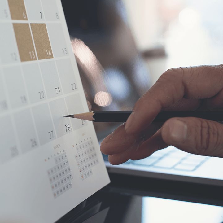 A hand holding a pencil points at a day on a desk calendar, a laptop behind it