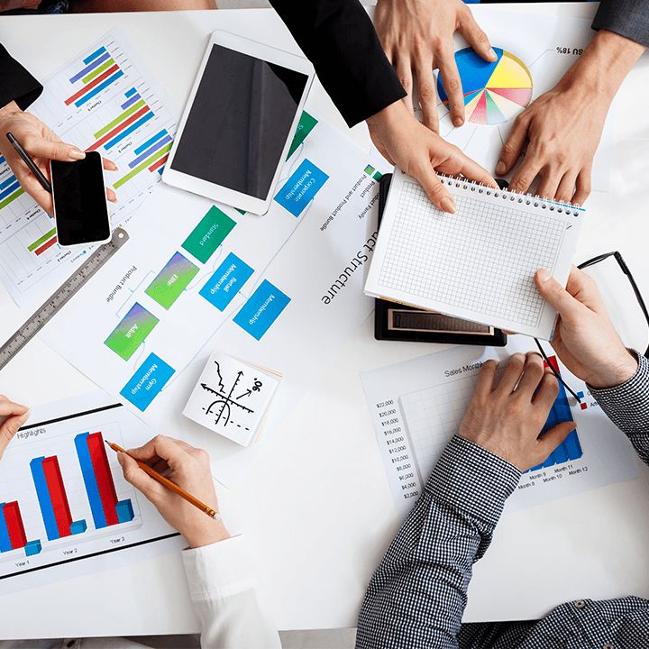 A boardroom table from above: hands at work over printed charts, a tablet and a notebook